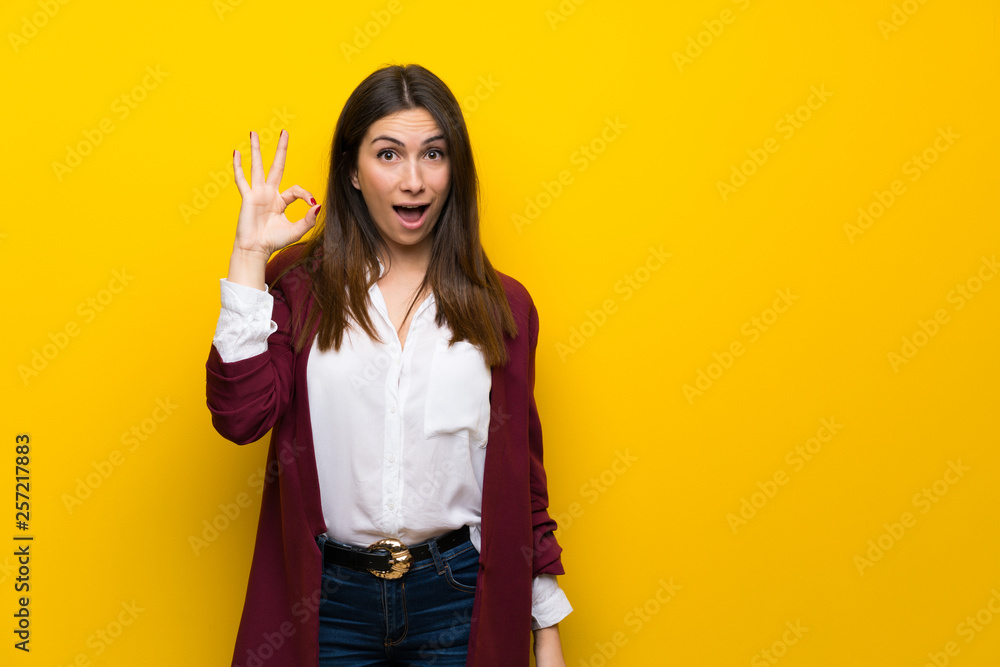 Young woman over yellow wall surprised and showing ok sign Young woman over yellow wall surprised and showing ok sign