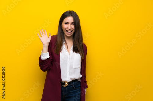 Young woman over yellow wall saluting with hand with happy expression
