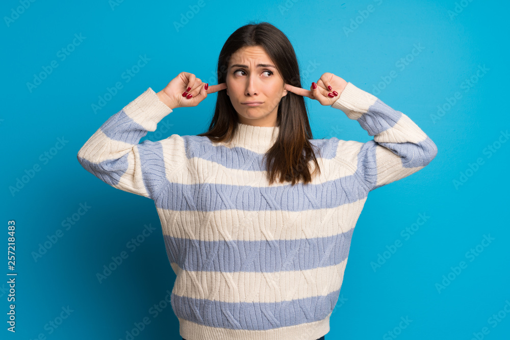 Young woman over blue wall covering both ears with hands Young woman over blue wall covering both ears with hands