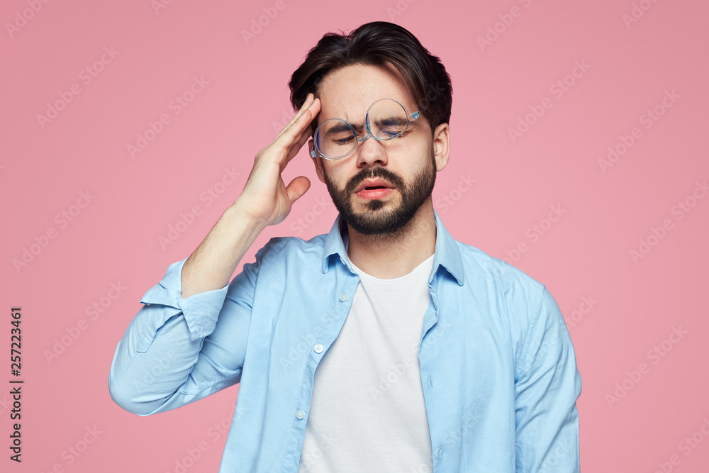 Portrait of tired exhausted young man with glasses, has terrible headache after work, being fed up with everything, closes eyes and keeps hands on temples as feels pain, isolated over pink background Portrait of tired exhausted young man with glasses, has terrible headache after work, being fed up with everything, closes eyes and keeps hands on temples as feels pain, isolated over pink background