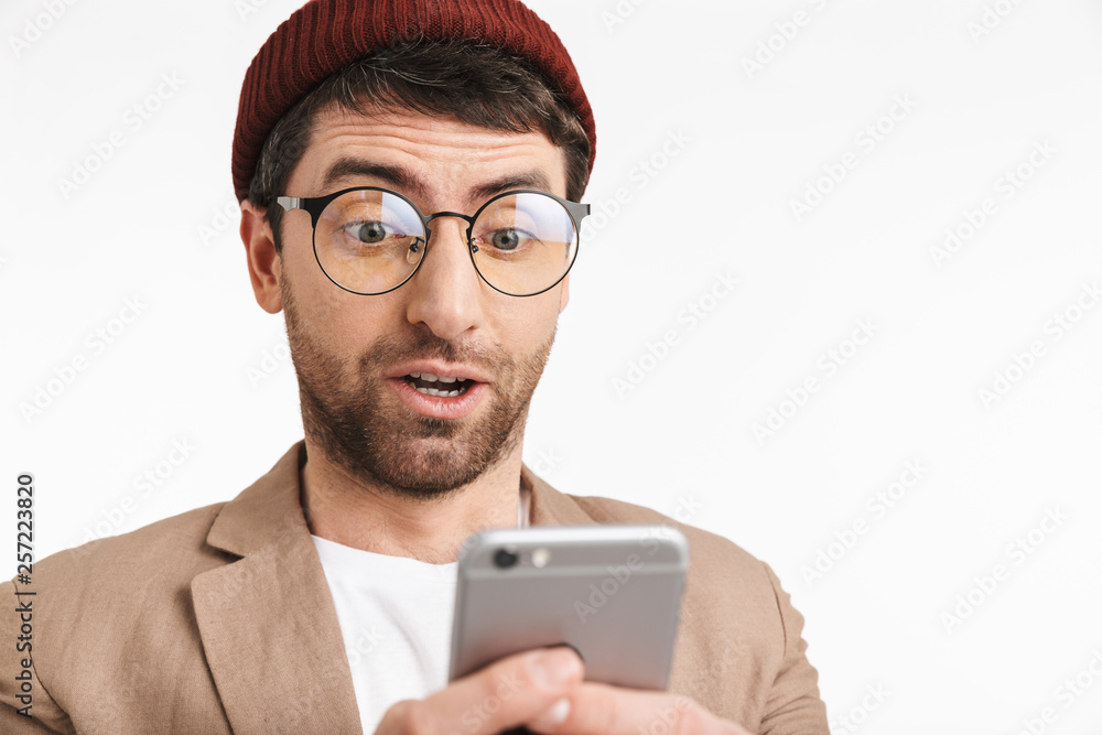 Photo of puzzled man 30s wearing hipster hat smiling while holding and using smartphone Photo of puzzled man 30s wearing hipster hat smiling while holding and using smartphone