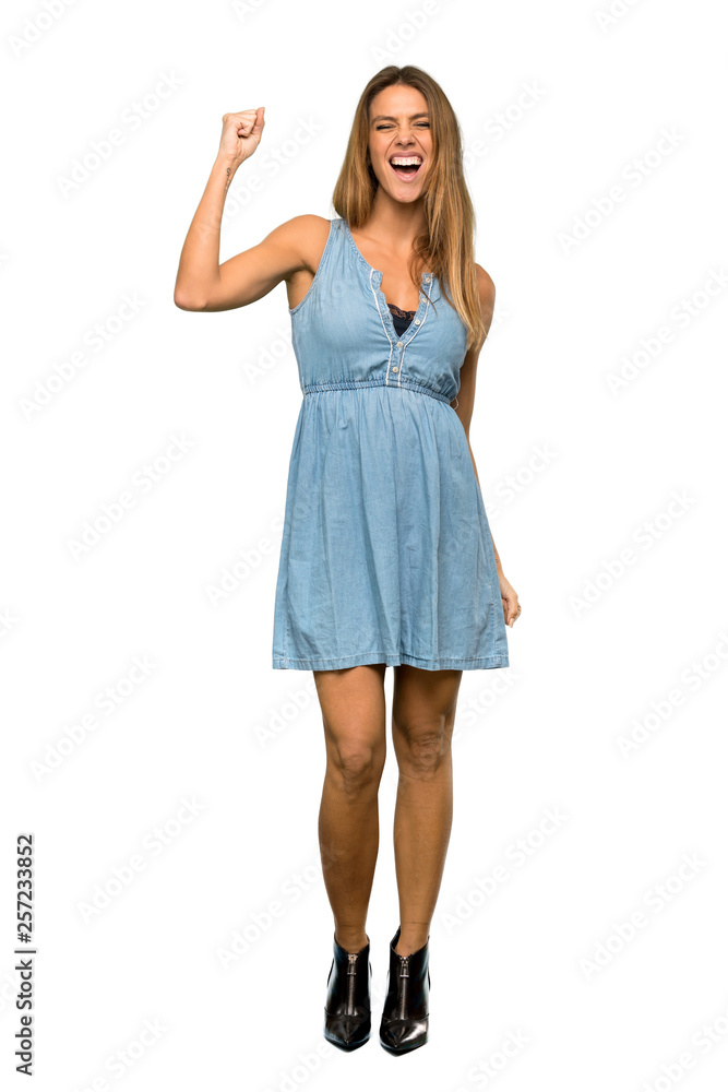 A full-length shot of a Blonde woman with jean dress celebrating a victory in winner position over isolated white background A full-length shot of a Blonde woman with jean dress celebrating a victory in winner position over isolated white background