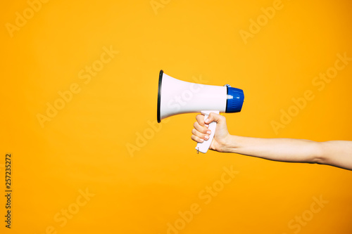 In a bull voice. A yellow background with a woman’s hand in front of it holding three colored megaphone of white, black and blue colors.