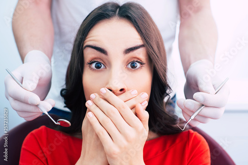 Foto Oh no! Plum scared of dentist work girl is hiding her mouth with the help of her hands while doctor is trying to do his work with a help of instruments and surgical gloves