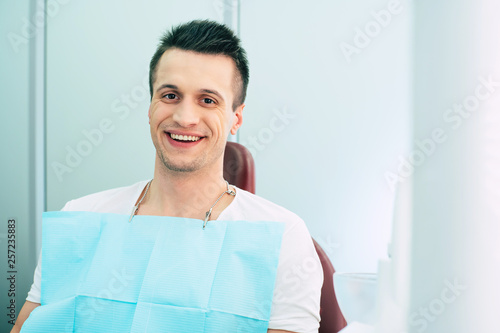 Gain point. A man with bodacious smile, hazel eyes and dark hair is sitting in a dental chair with a special napkin over his neck and smiling right into the camera. photo