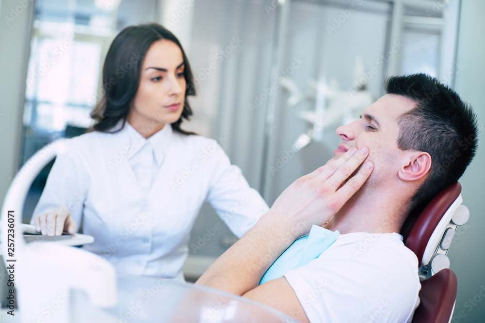 In need of a medical help. A man in a dental chair near the professional dentist is showing that he has a horrible tooth pain with his gestures and asking for a treatment. In need of a medical help. A man in a dental chair near the professional dentist is showing that he has a horrible tooth pain with his gestures and asking for a treatment.