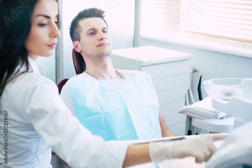 Treatment session. A man who came in a dental clinic to have a treatment is sitting in a dental chair near the dentist who is getting ready to start working in the problem of a patient.