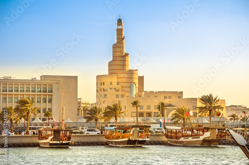 Fotografia Traditional wooden dhow anchored at Dhow Harbor in Doha Bay with spiral mosque and minaret in the background at sunset