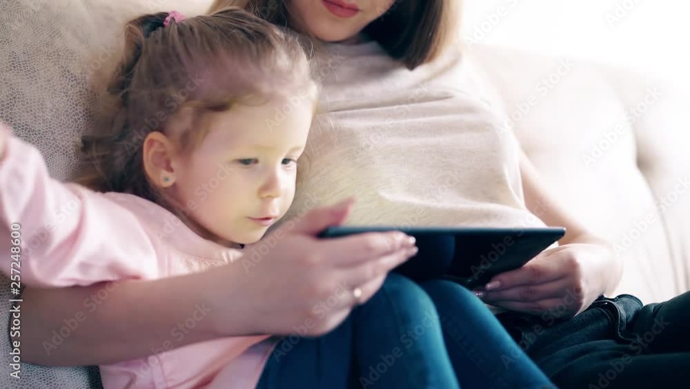 CU: Side view, 4k: Young attractive mother and sweet daughter are sitting on the couch and teaching using a tablet computer. The daughter passes her fingers along the screen. Rejoices. Smiling. CU: Side view, 4k: Young attractive mother and sweet daughter are sitting on the couch and teaching using a tablet computer. The daughter passes her fingers along the screen. Rejoices. Smiling.