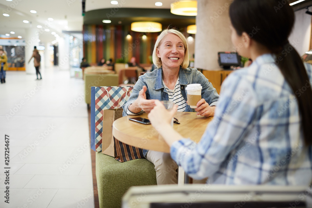 Happy excited mature woman with blond hair sitting at table and gesturing hand while telling story to friend and drinking coffee in food court of shopping mall.