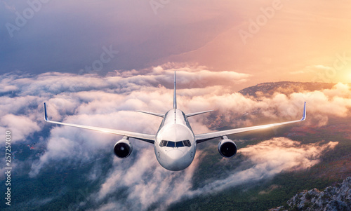 Airplane is flying over clouds at sunset in summer. Landscape with passenger airplane, low clouds, sea, purple sky at dusk. Front view of the aircraft. Business travel. Commercial plane. Aerial view