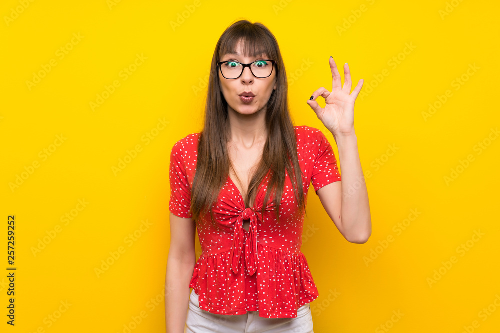 Young woman over yellow wall surprised and showing ok sign Young woman over yellow wall surprised and showing ok sign