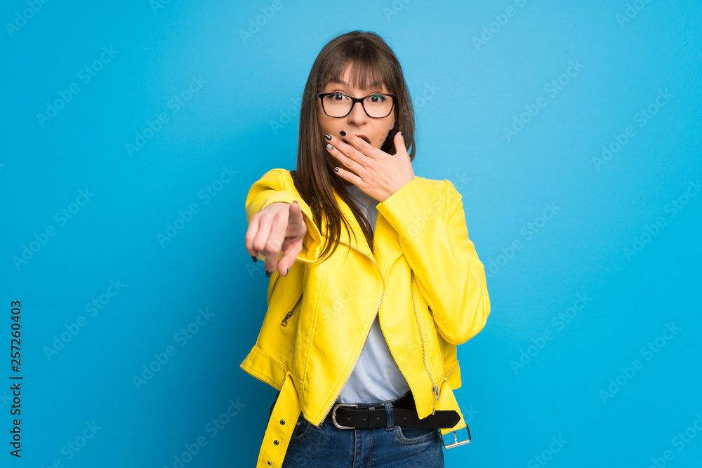 Young woman with yellow jacket on blue background pointing with finger at someone and laughing Young woman with yellow jacket on blue background pointing with finger at someone and laughing