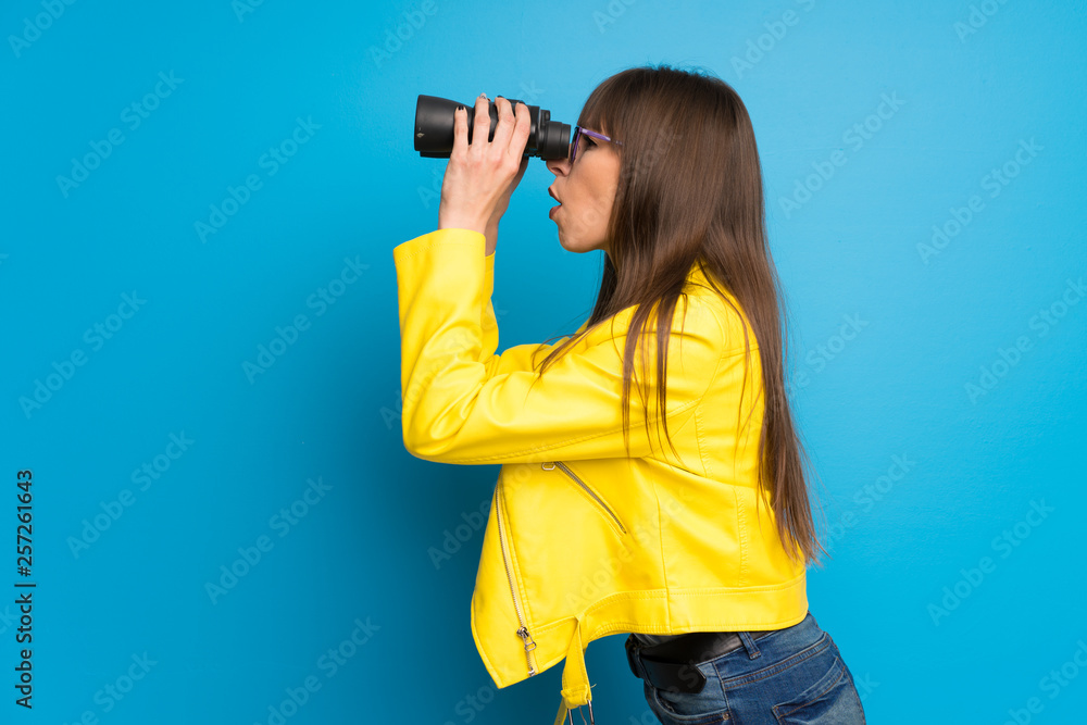 Young woman with yellow jacket on blue background and looking in the distance with binoculars Young woman with yellow jacket on blue background and looking in the distance with binoculars
