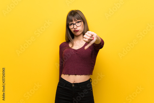 Woman with glasses over yellow wall showing thumb down with negative expression