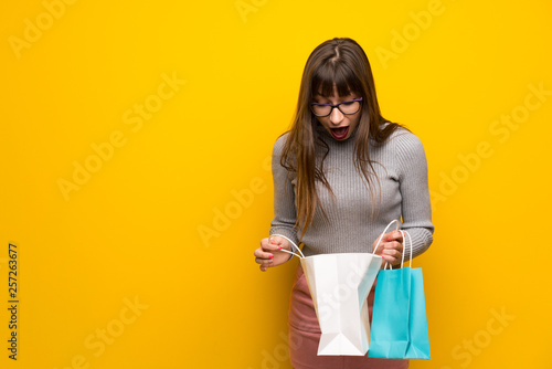 Woman with glasses over yellow wall surprised while holding a lot of shopping bags © luismolinero