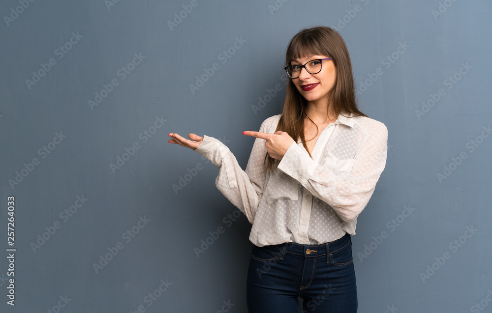 Woman with glasses over blue wall holding copyspace imaginary on the palm to insert an ad Woman with glasses over blue wall holding copyspace imaginary on the palm to insert an ad