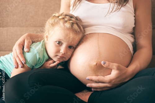 Close-up view of lovely girl sitting on sofa with pregnant mother and looking at camera. Cheerful woman embracing daughter and expecting baby. Concept of parental love and pregnancy.