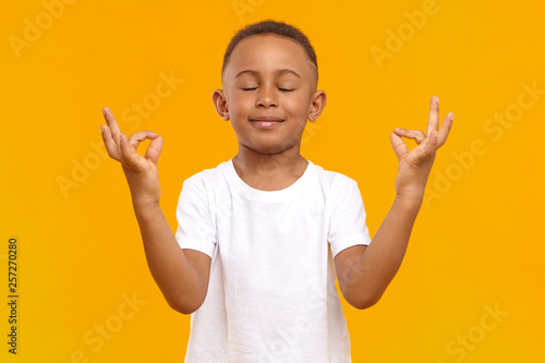 Quadro su tela Peaceful calm little boy of African appearance wearing casual t-shirt keeping his eyes closed and smiling, meditating isolated in studio