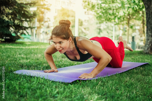  a young girl on the street in a city park on a lawn doing exercises, doing yoga and doing stretching. On the girl red leggings