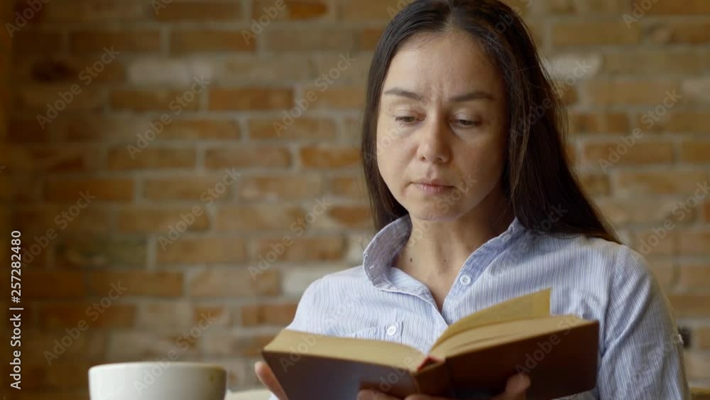 serious middle-aged brunette woman sitting in a cafe against a brick wall, reading a book and drinking coffee. serious middle-aged brunette woman sitting in a cafe against a brick wall, reading a book and drinking coffee.