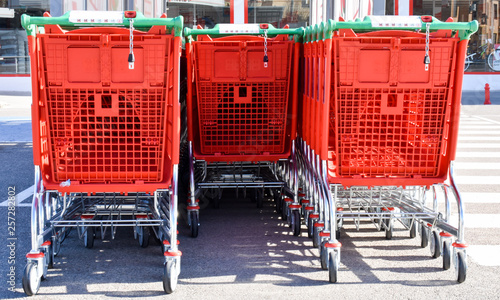 close up of red metallic and plastic trolleys tidied in several rows waiting for being used by buyers at a parking place in a supermarket. Horizontal