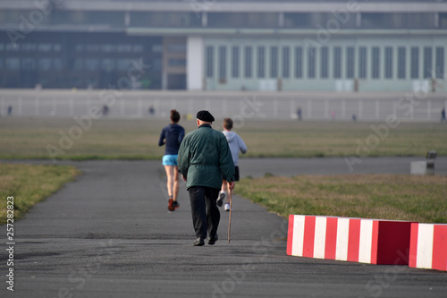 An elderly man with a black hat is walking near the airport runway of the airport Berlin-Sch  nefeld. Young men walking at some distance in front of an elderly man on a warm spring day.
