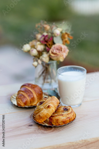 Buns with raisins and french croissant. A glass of milk on rustic table. Dinner in an orchard in summertime. Bouquet of flowers. Dessert. Soft focus. Blurry background. Copy space.