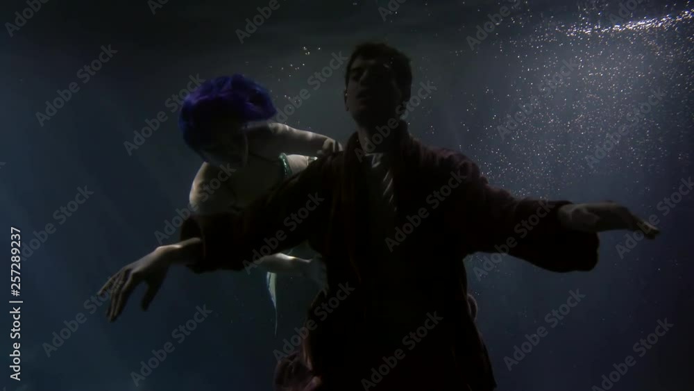 A young man with his eyes closed, slowly falling under the water and circling around him, swims a beautiful mermaid girl with blue hair A young man with his eyes closed, slowly falling under the water and circling around him, swims a beautiful mermaid girl with blue hair