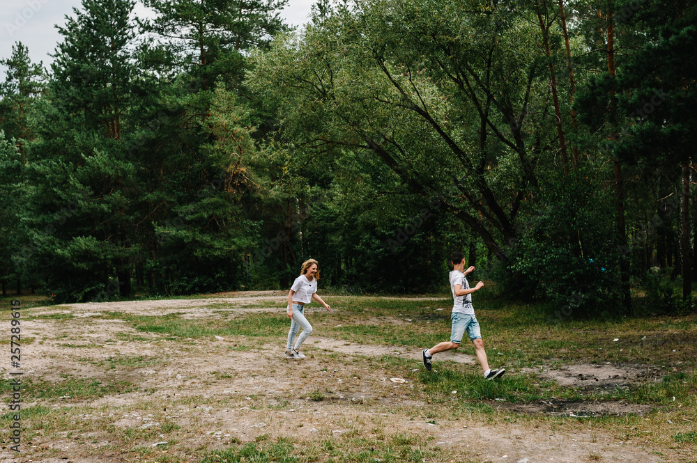 Fototapeta premium Young couple running away and having fun on the background on the nature. A woman catches her husband against the background of trees and fir trees.