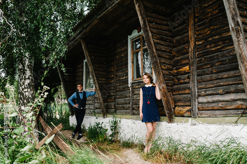 Young couple standing near an old wooden stylish house on the background of birch and nature. A man is far from a woman. full length. looking sideways  at camera.