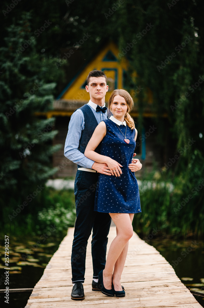 Φωτογραφία The young couple standing on a wooden bridge above the lake on the background a old house on an island  near of trees