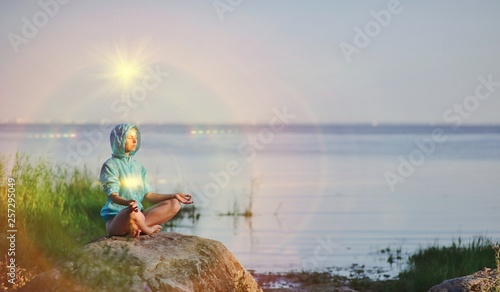 Beautiful peaceful woman sits in a pose of a half lotus practicing yoga meditation, blue summer sky, glowing chakra, eyes closed calm. Kundalini energy exercise. Human spirit, people healing concept