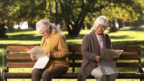 Two senior offended female friends reading newspapers, sitting on bench in park