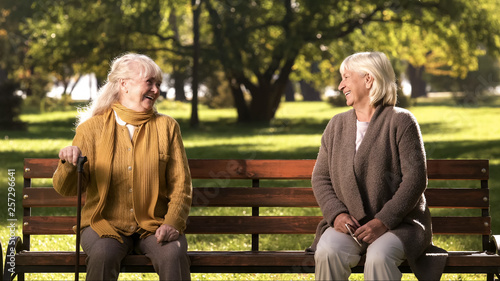 Two elderly ladies laughing and talking, sitting on bench in park, old friends