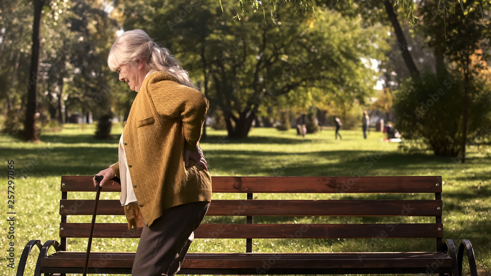 Fototapeta premium Senior lady slowly walking with stick along park suffering from back pain