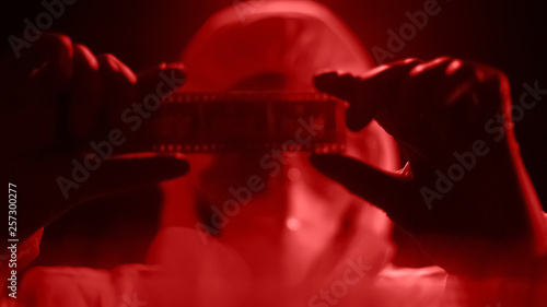 Принти на полотні Man in lab costume watching camera roll, developing photograph in red light