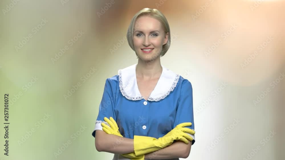 Portrait of smiling young housemaid with arms crossed. Cheerful cleaner woman in blue uniform and yellow gloves looking at camera. Abstract blurred background. Portrait of smiling young housemaid with arms crossed. Cheerful cleaner woman in blue uniform and yellow gloves looking at camera. Abstract blurred background.