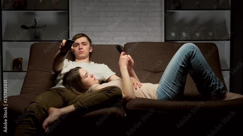 Tired man switches channels on TV while using the remote control sitting on the sofa. girl lies with the phone. Tired man switches channels on TV while using the remote control sitting on the sofa. girl lies with the phone.