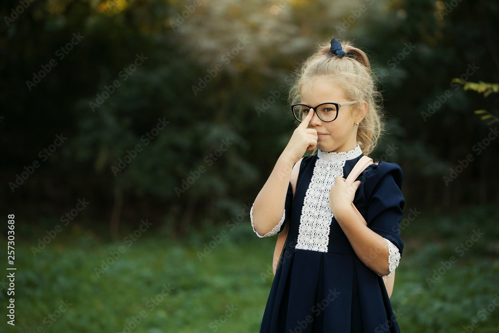 Little happy smiling smart girl in glasses and uniform standing in the backyard of the school. schoolgirl goes to first grade. Copy space for text on left side Little happy smiling smart girl in glasses and uniform standing in the backyard of the school. schoolgirl goes to first grade. Copy space for text on left side