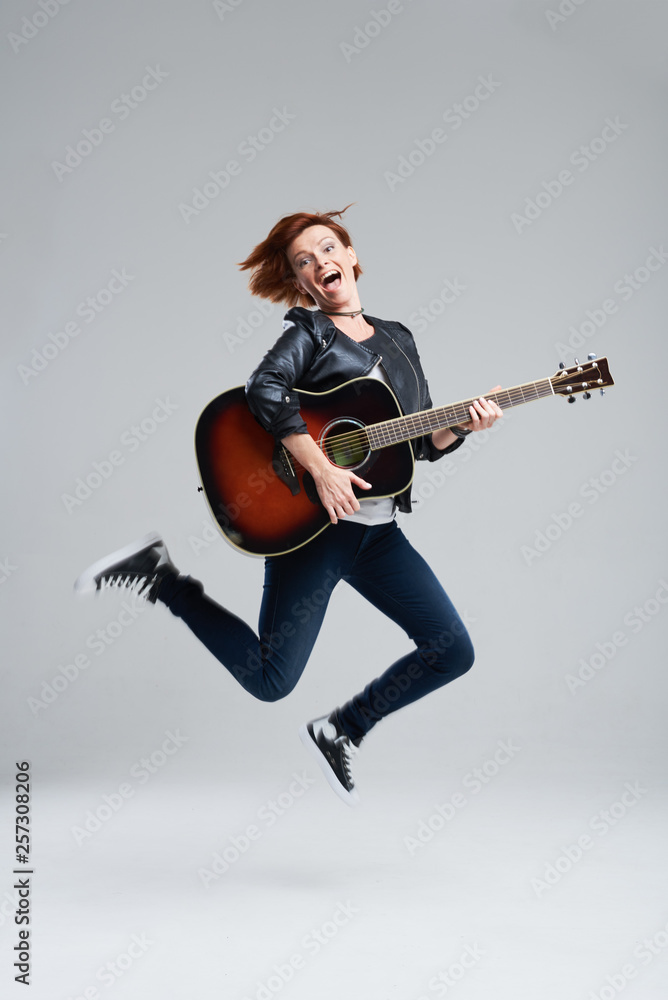 Young woman musician with an acoustic guitar in hand on a gray background. She laughs and jumps high. plays rock and roll loudly. Full-length portrait. 