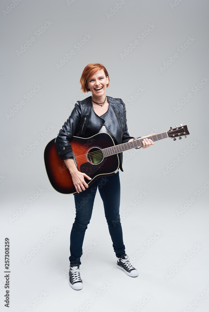 Young woman musician with an acoustic guitar in hand on a gray background. She plays rock and roll loudly. Full-length portrait. 