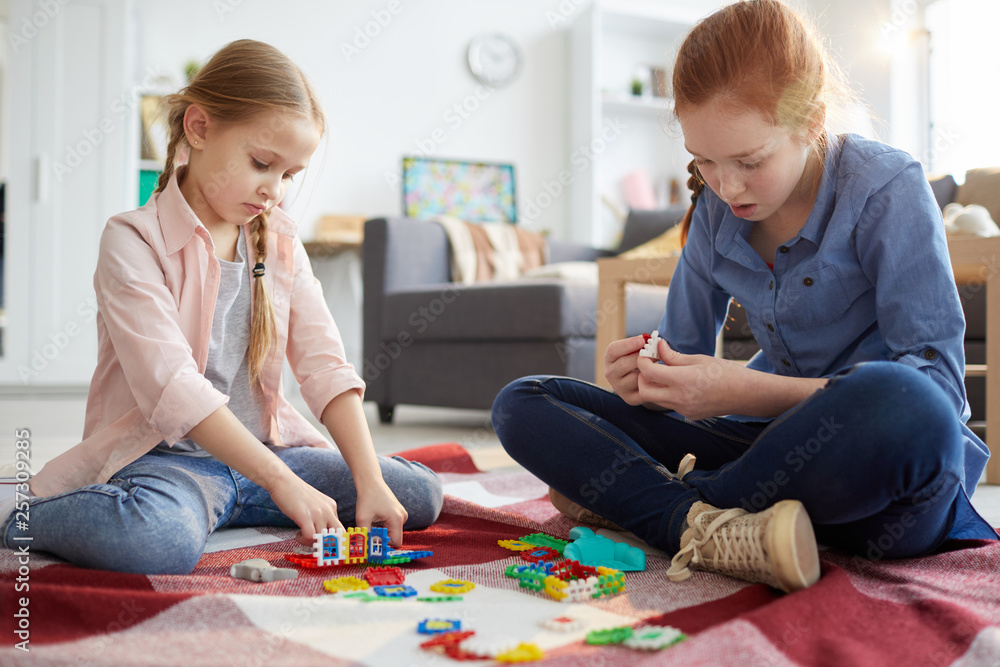 Fototapeta premium Full length portrait of two sisters playing with puzzles sitting on floor at home, copy space