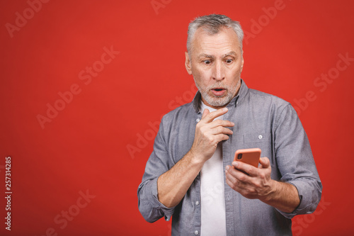 Portrait of shocked senior man in casual gasping opening mouth from worry and surprise holding mobile phone, looking in camera isolated over red background. © denis_vermenko
