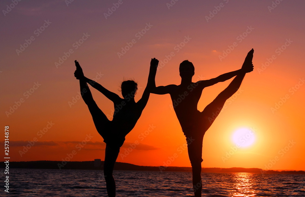 A pair of gymnasts athletes perform in nature on a sunset background. Silhouette photo of two people exercising against the sea raising their legs up and sitting on the splits. The girl bends the leg 