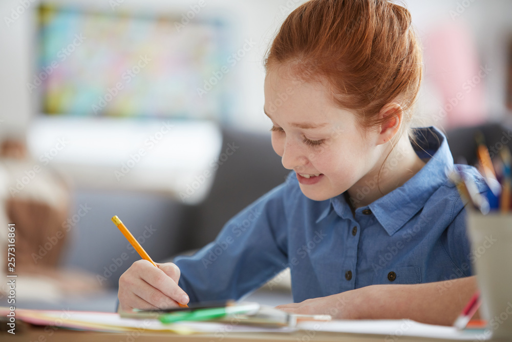 Portrait of red-haired schoolgirl studying sitting at desk and writing with pencil, copy space Portrait of red-haired schoolgirl studying sitting at desk and writing with pencil, copy space