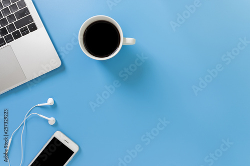 Listening to music at work. Flat lay desk top with laptop, earphone, coffee on blue background. Cool tone.