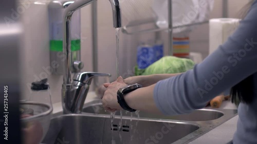 Young woman washing her hands and using her smartwatch in kitchen at night. Camera on slider