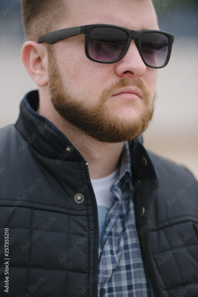 A man with a beard and sunglasses in a plaid shirt and waistcoat posing on the street to advertise men's clothing. Advertising menswear A man with a beard and sunglasses in a plaid shirt and waistcoat posing on the street to advertise men's clothing. Advertising menswear
