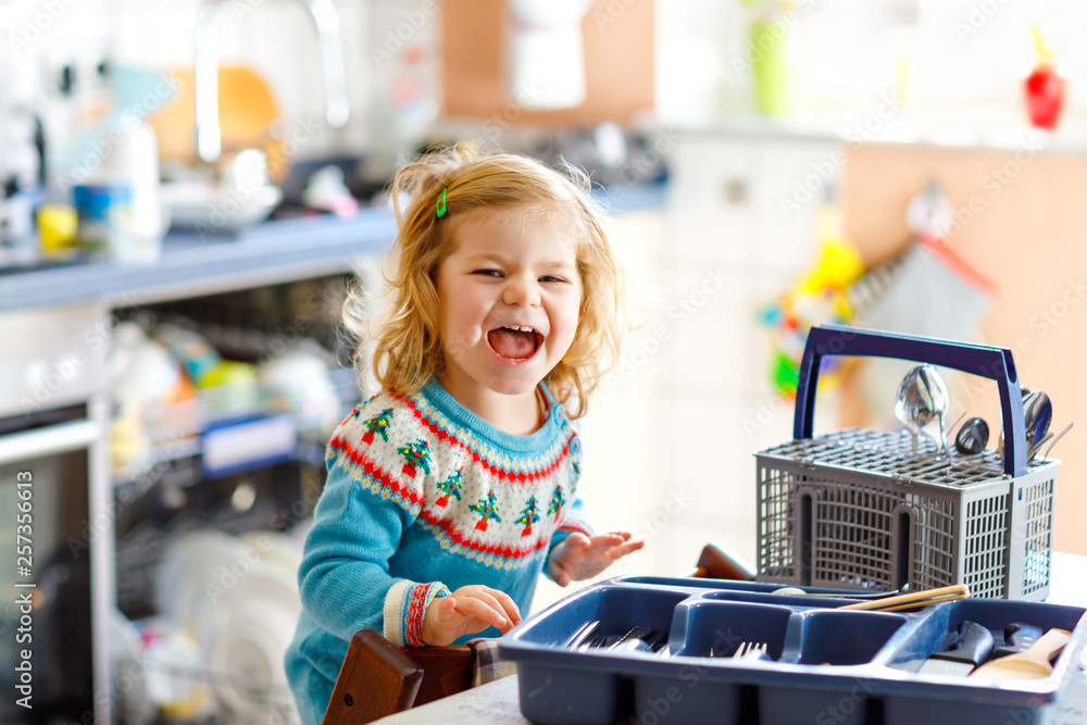 Cute little toddler girl helping in the kitchen with dish washing machine. Happy healthy blonde child sorting knives, forks, spoons, cutlery. Baby having fun with helping housework mother and father. Cute little toddler girl helping in the kitchen with dish washing machine. Happy healthy blonde child sorting knives, forks, spoons, cutlery. Baby having fun with helping housework mother and father.
